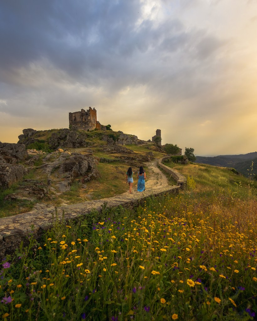 Dos chicas pasean al atardecer con un castillo en ruinas de fondo y flores en primer plano